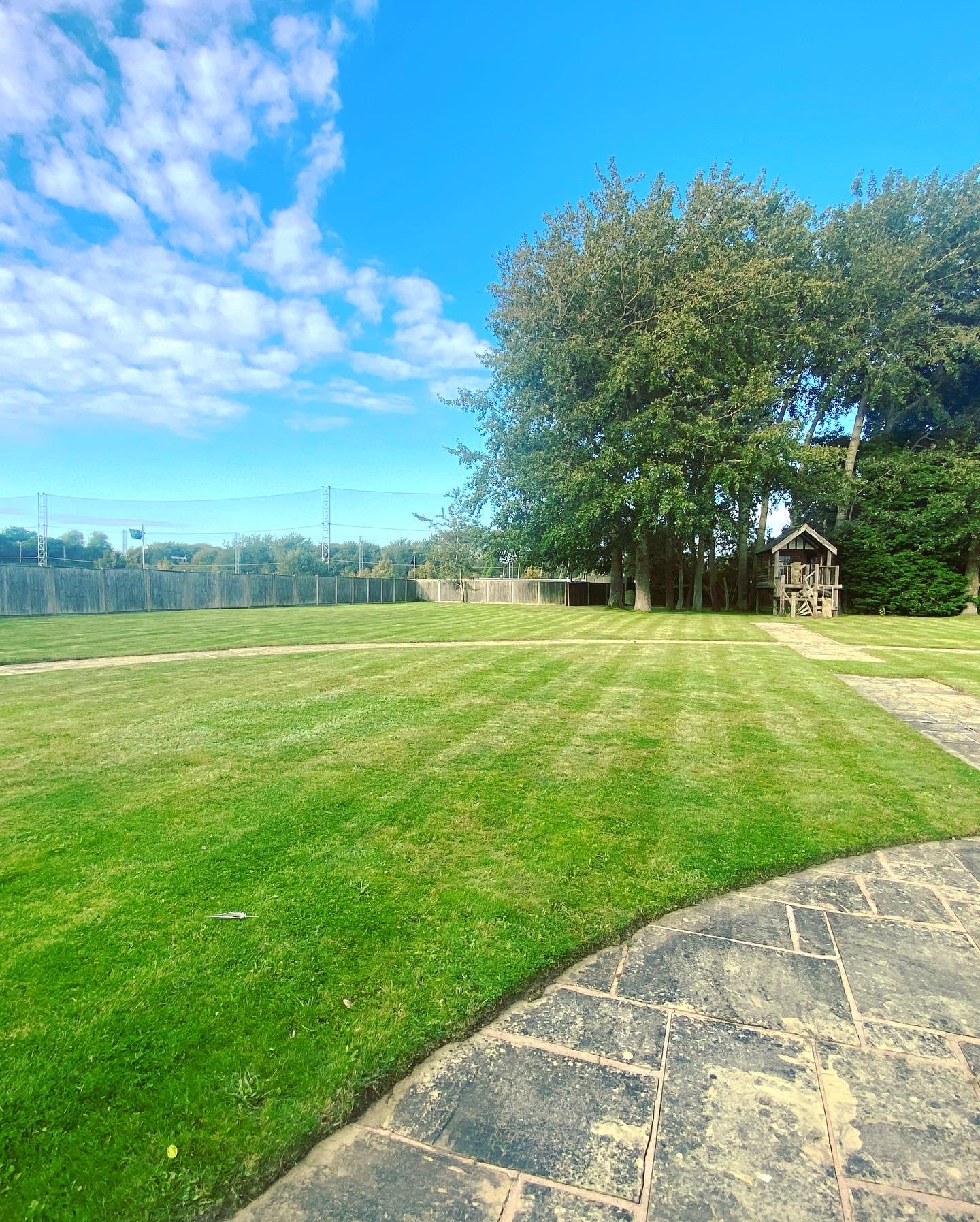 A backyard with a well-maintained grassy lawn, a stone pathway, a wooden playhouse near tall green trees, a wooden fence, and a partly cloudy sky.