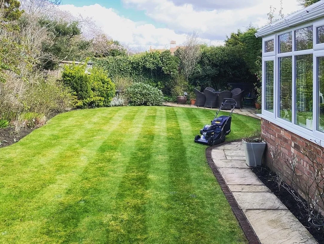 A well-maintained backyard with striped green grass, edged with a stone walkway, containing potted plants and a lawn mower, with a patio area furnished with black outdoor furniture, and surrounded by tall bushes and trees.