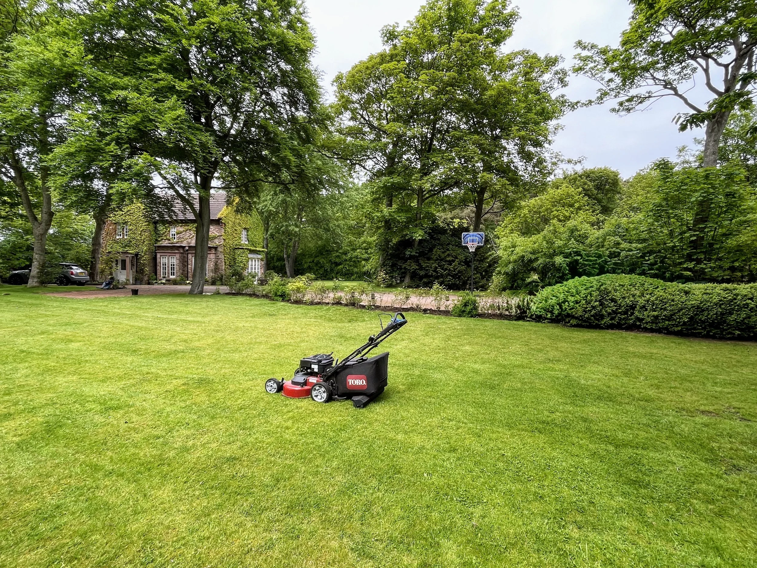 A pristine, well-maintained backyard featuring lush green grass, tall leafy trees, and a brick house with ivy climbing its walls. In the yard, a red and black Toro lawn mower is positioned on the grass, and a black lawn chair is nearby. In the background, a basketball hoop is set up, and a parked car is visible near the house.