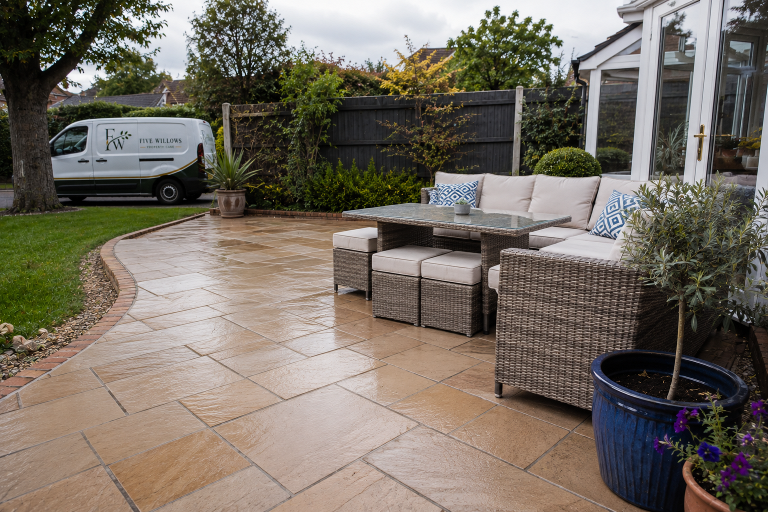 A wet outdoor patio with patio furniture, including a sectional sofa, a table, and stools. There are potted plants and trees, a garden, and a white van with the logo 'Five Willows Property Care' parked nearby. The scene is overcast, and the patio surface is wet.