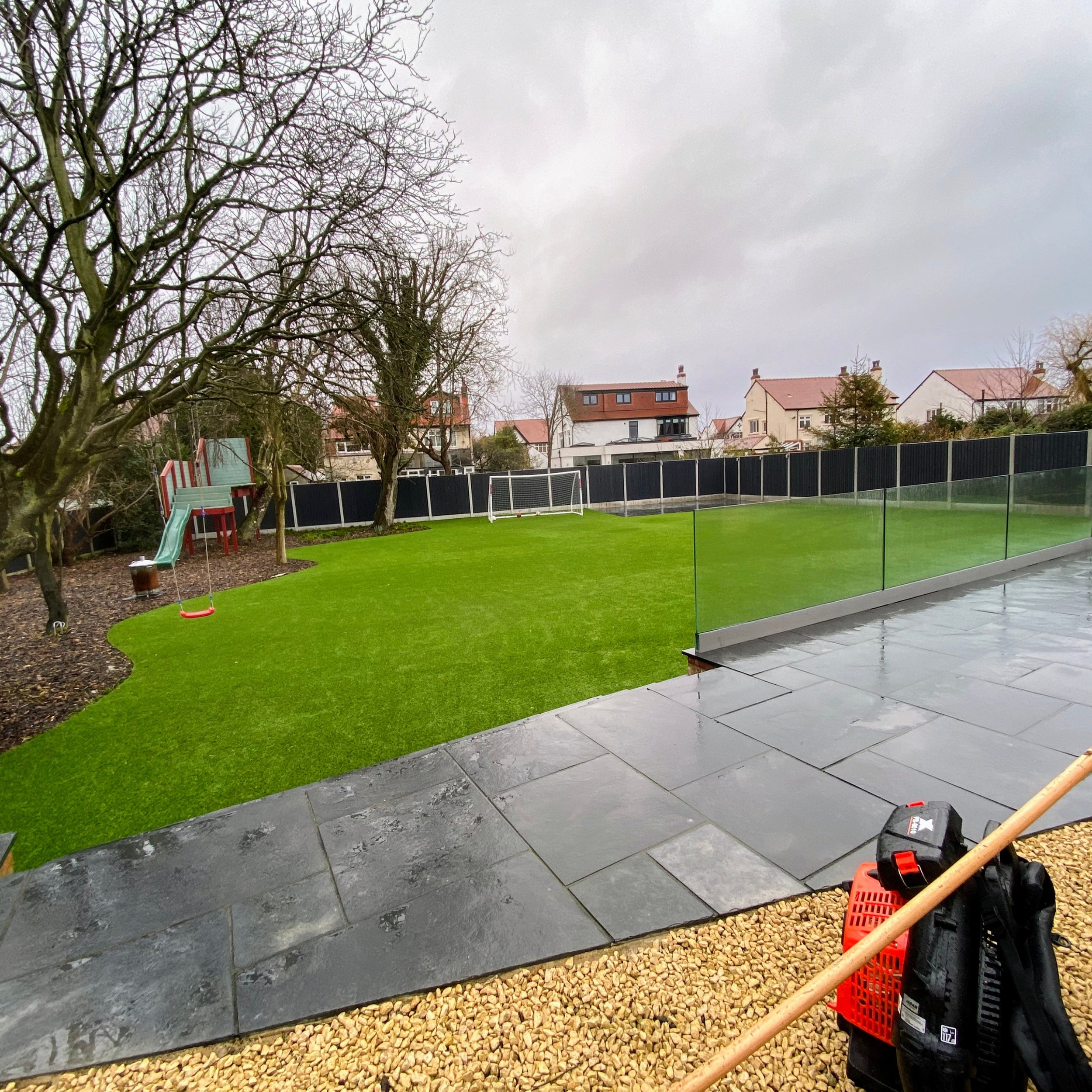 A backyard with a neatly mowed green lawn, a small slide, and a soccer goal. There are leafless trees, a black fence, and houses with red roofs in the background. The sky is overcast.