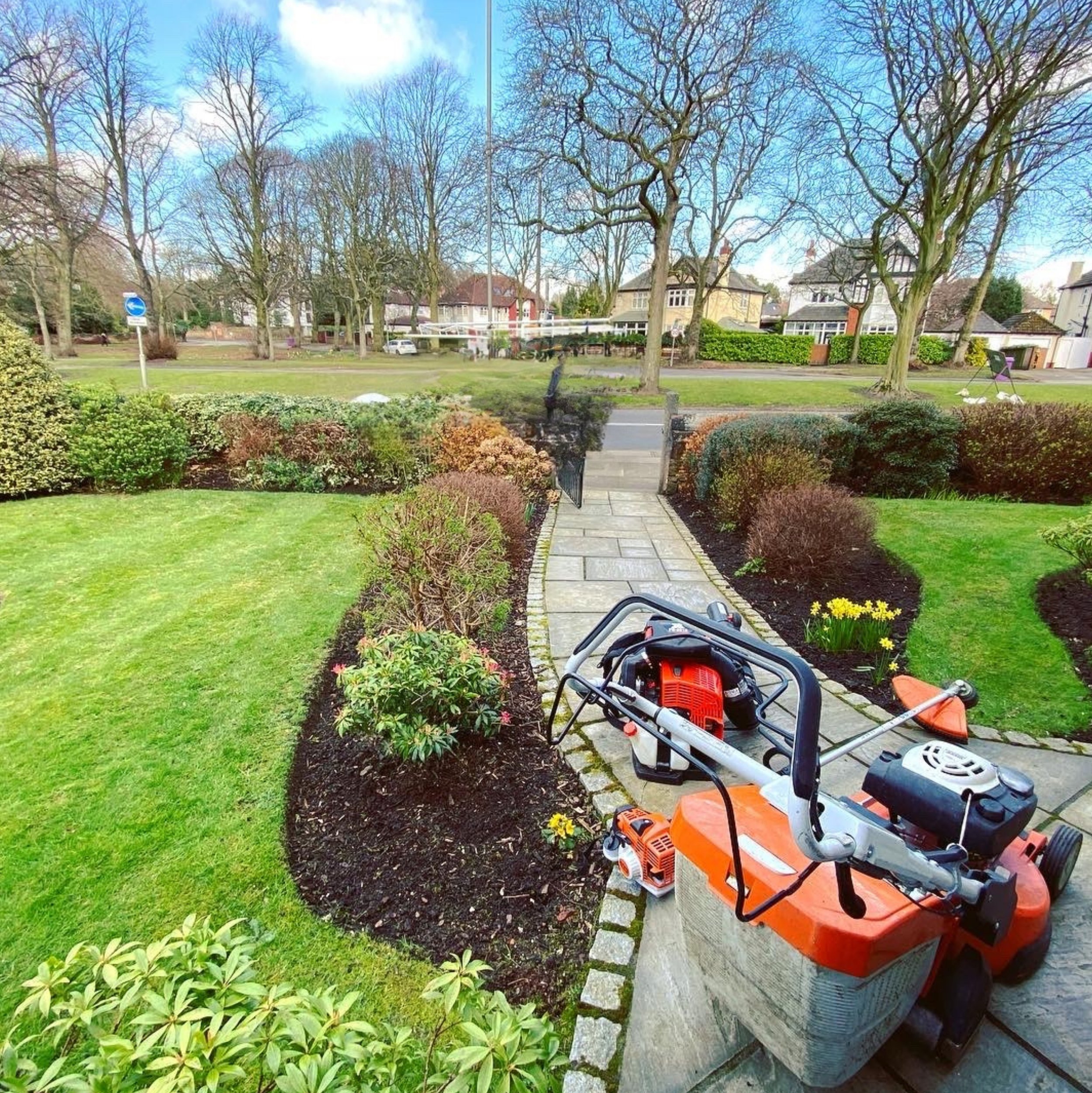 A garden with a paved pathway, green lawn, and flower bed near the edge. Garden tools and equipment, including a leaf blower, are placed on a bench beside the path. The background features leafless trees, houses, and a partly cloudy sky.