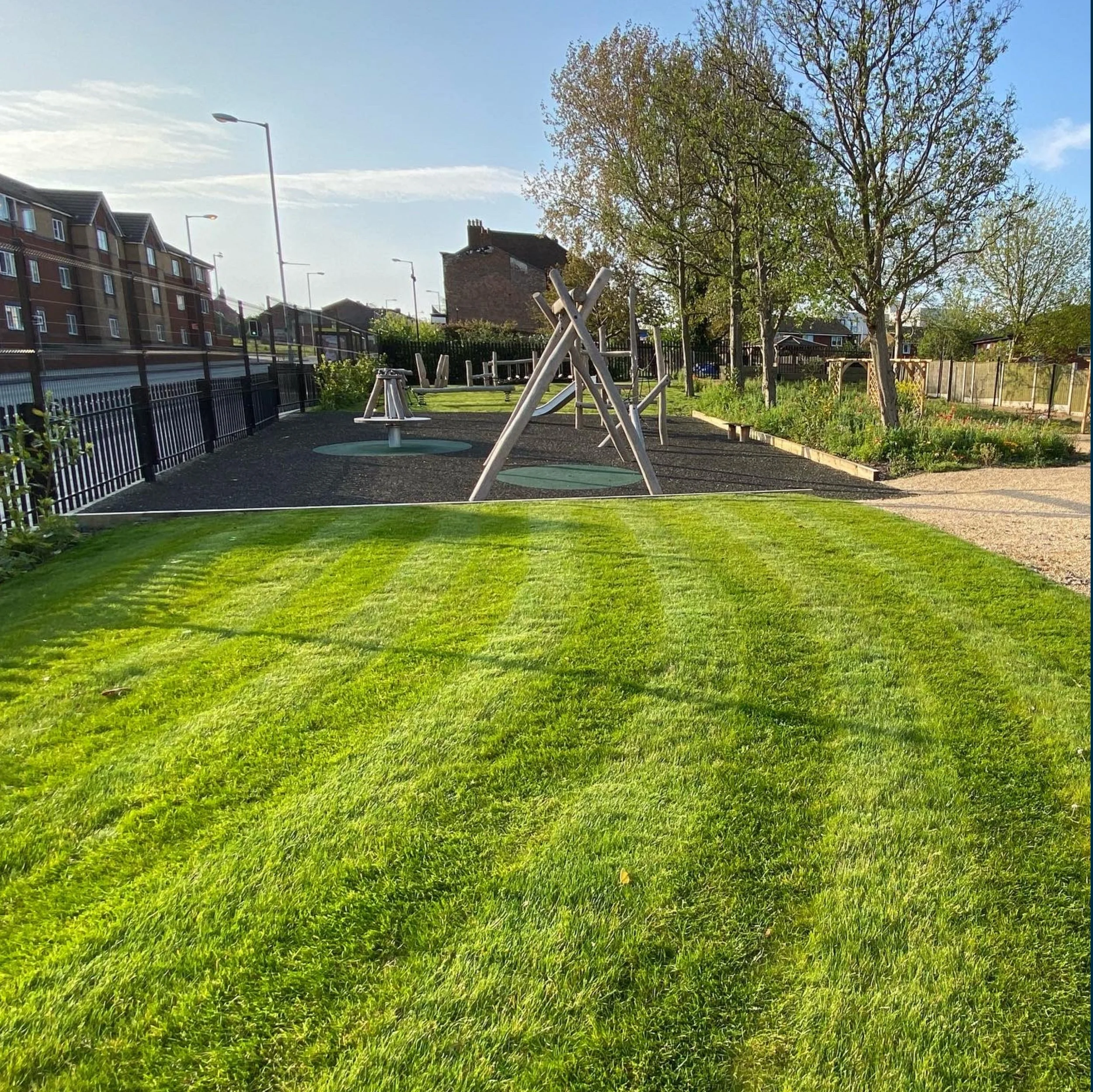 A playground with white climbing frames and a green grassy field, surrounded by trees and residential buildings, bathed in sunlight on a clear day.