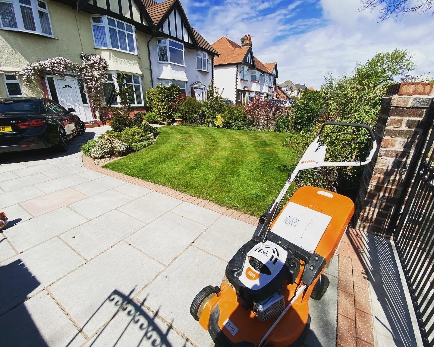 A front yard with a small lawn, flower beds, a driveway with a black car, and a leaf blower machine in the foreground. Houses with tiled roofs and a partly cloudy sky are in the background.