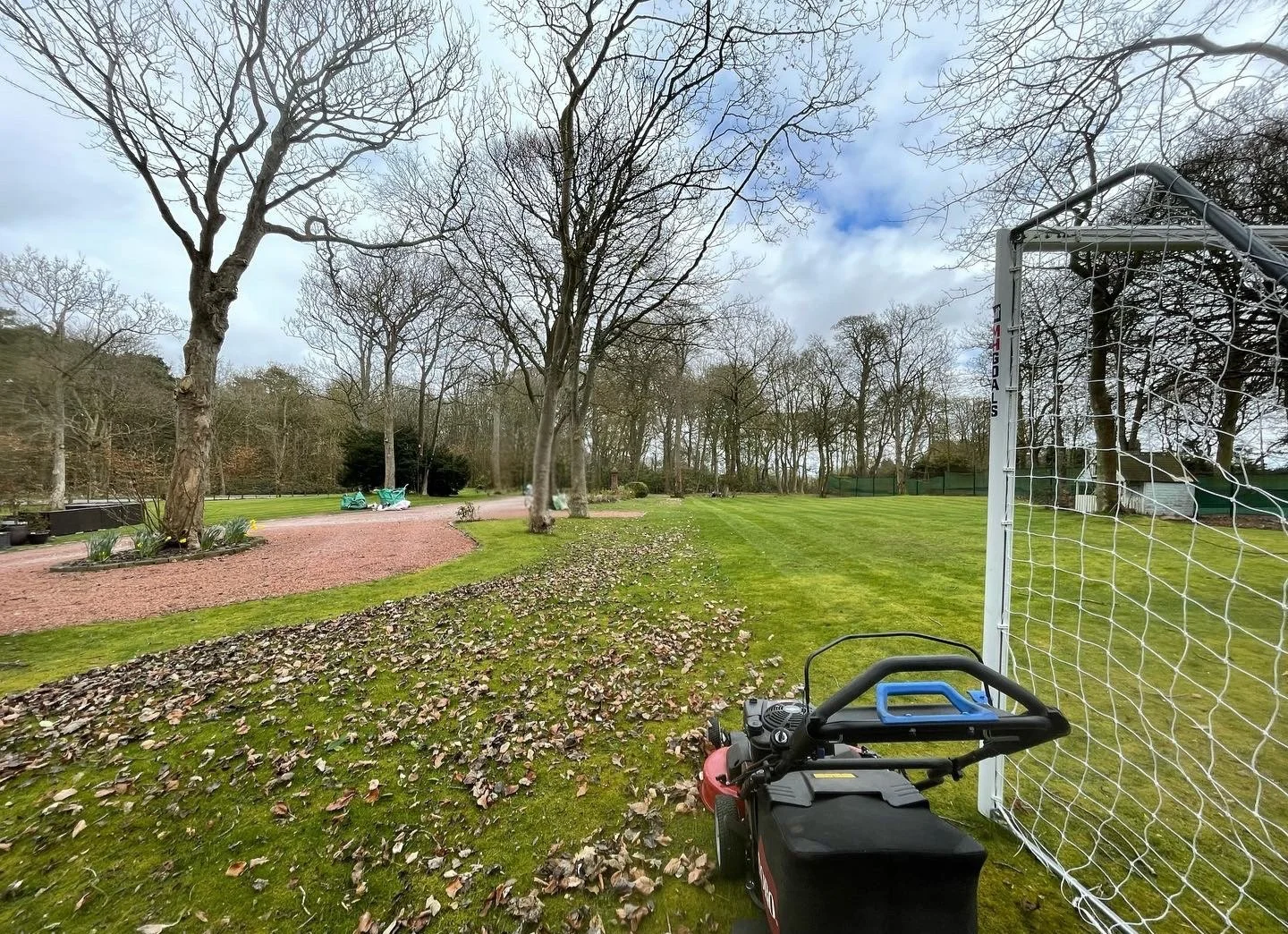 A well-maintained park with a grassy field, leafless trees, a gravel pathway, and a soccer goal on the right. A leaf blower is visible in the foreground, and the sky is partly cloudy.