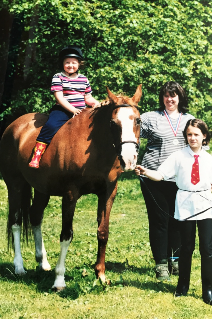 A young girl in colorful rain boots and a striped shirt is sitting on a brown horse with a white blaze on its face, while two older girls, one wearing a medal, stand next to the horse holding its reins outdoors on lush green grass with trees in the background.
