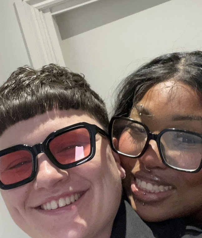 Two friends smiling and taking a close-up selfie indoors. One is wearing pink-tinted sunglasses, and the other has large glasses and piercings.