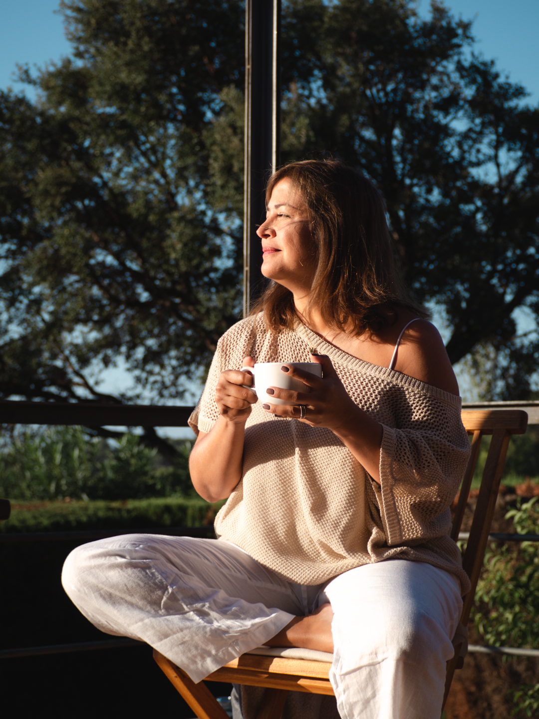 Mujer sentada en una silla de madera en un balcón, sosteniendo una taza, disfrutando del sol a través de un ventanal grande con árboles en el fondo.