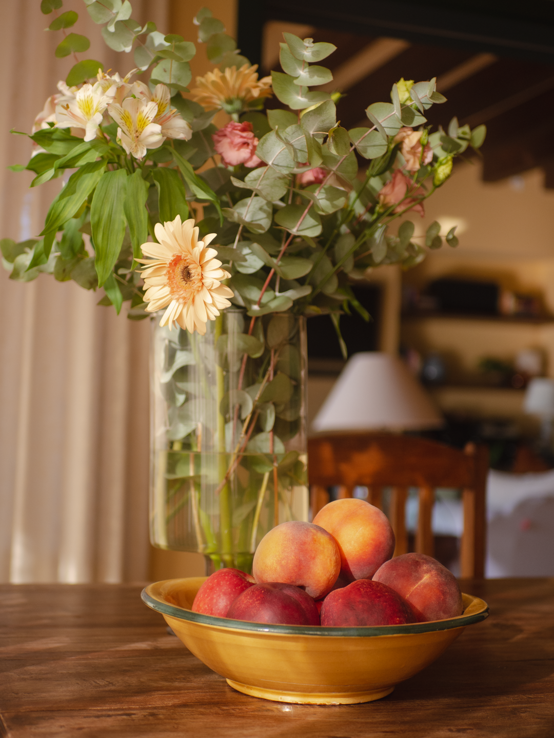 Florero con flores variadas y un plato con duraznos y manzanas en una mesa de madera en un ambiente acogedor.