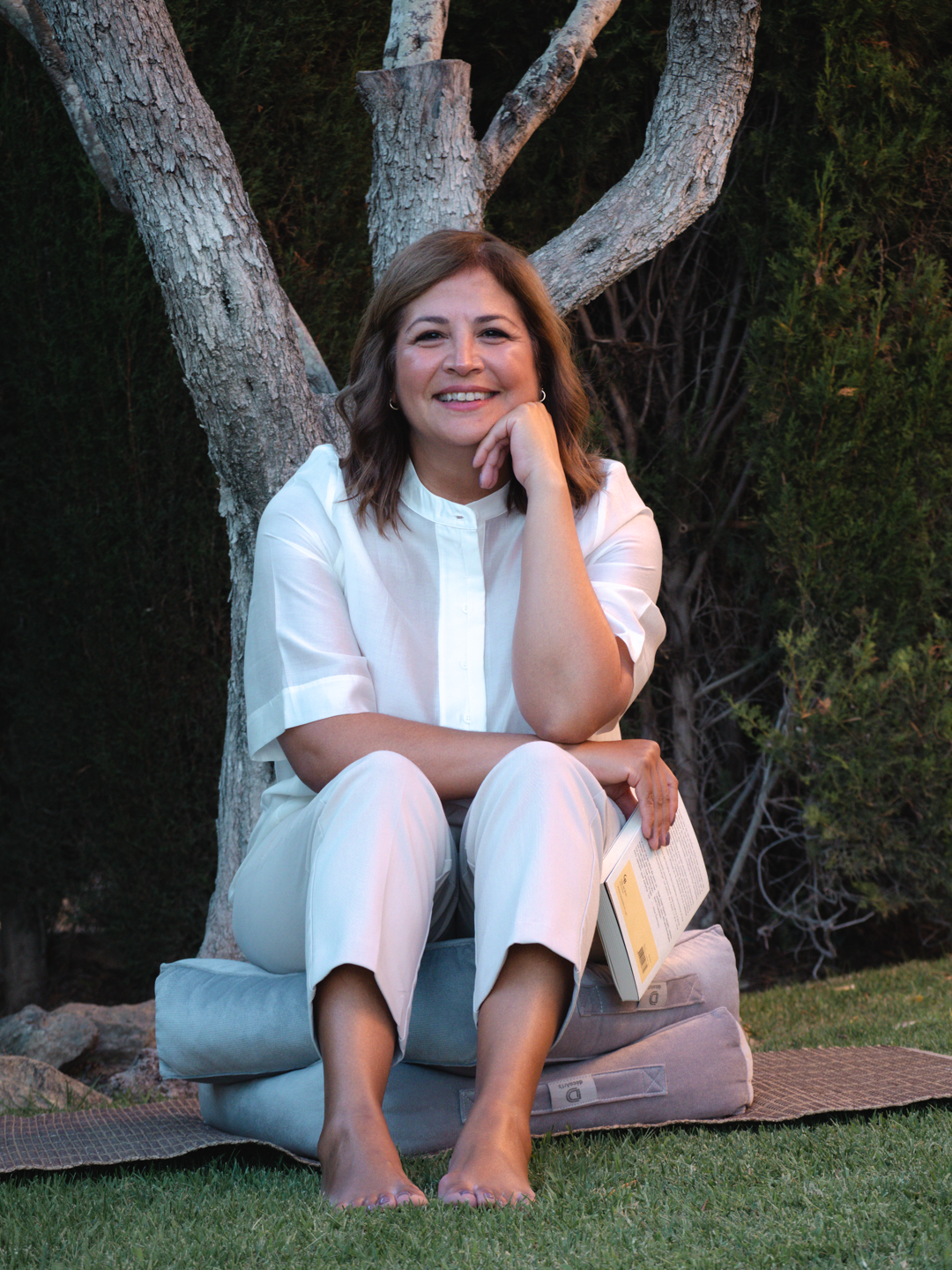 Mujer sonriendo, sentada en alfombra en un parque, con árboles y arbustos en el fondo, leyendo un libro.