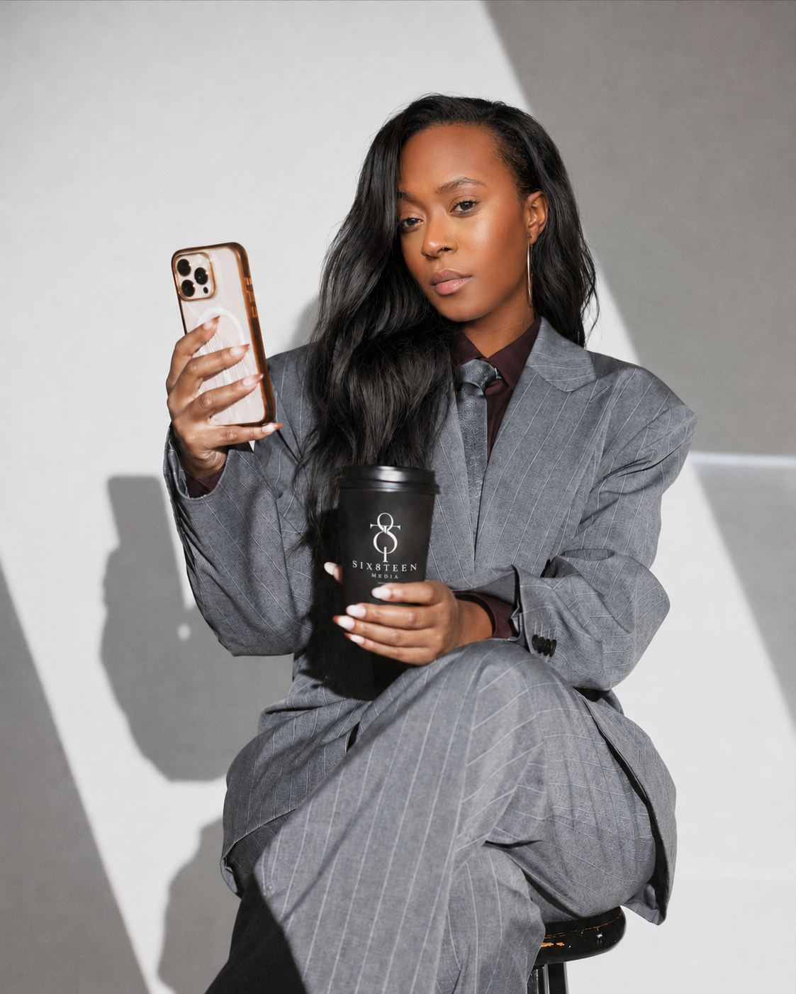 A woman in a gray striped business suit sitting on a stool, holding a black coffee cup labeled 'SIXTEEN MEDIA' in one hand and looking at her phone in the other.