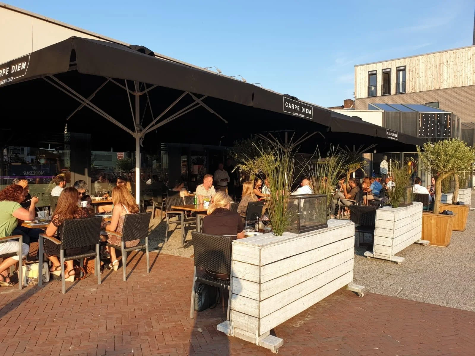 Buiten restaurant met grote zwarte parasols en tafels vol mensen, omgeven door planten en moderne gebouwen.