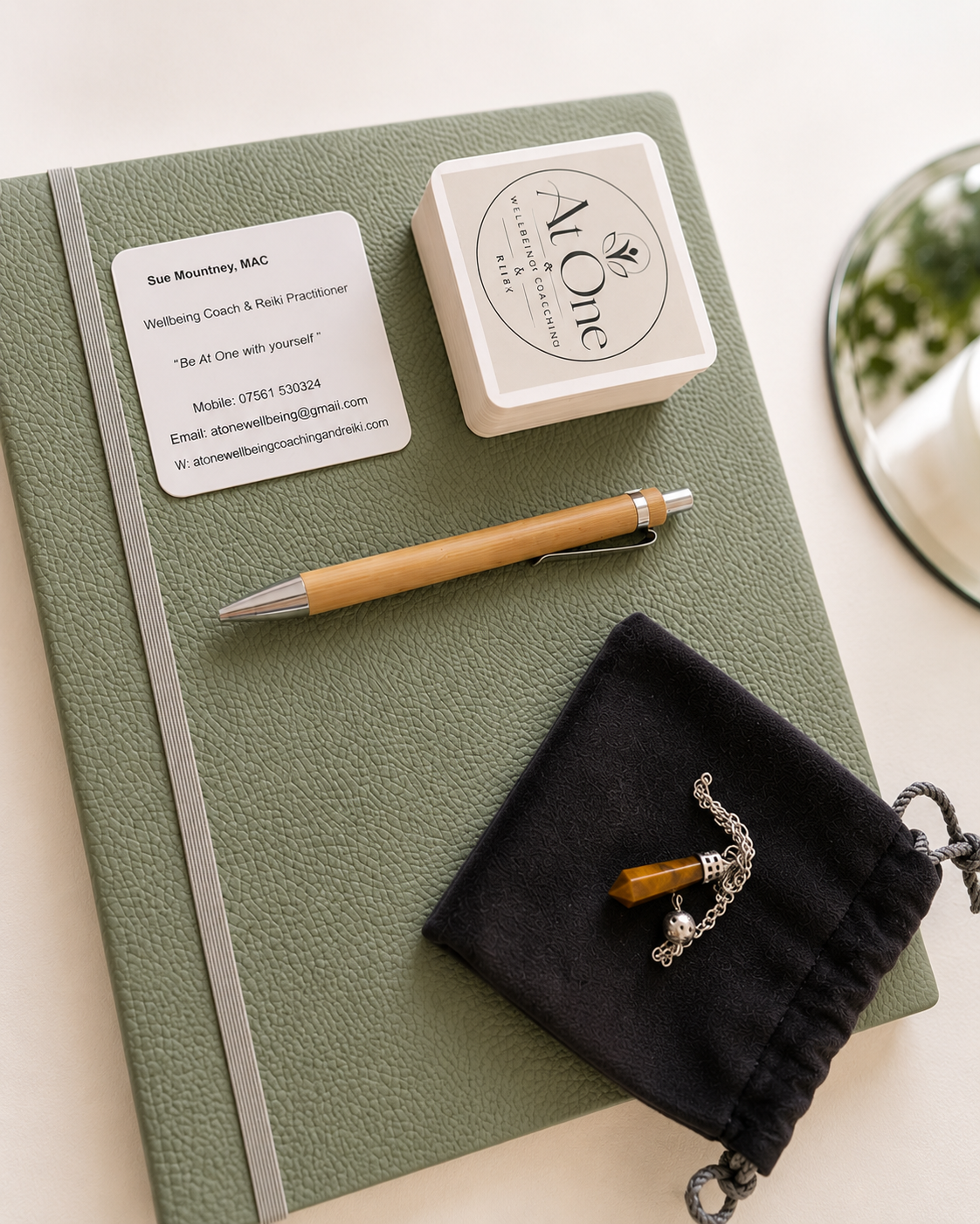 A green textured notebook with a business card, a deck of cards, a wooden pen, and a small black pouch with a chain bracelet on top. The business card belongs to Sue Mountney, a Wellbeing Coach and Reiki Practitioner, with contact details.