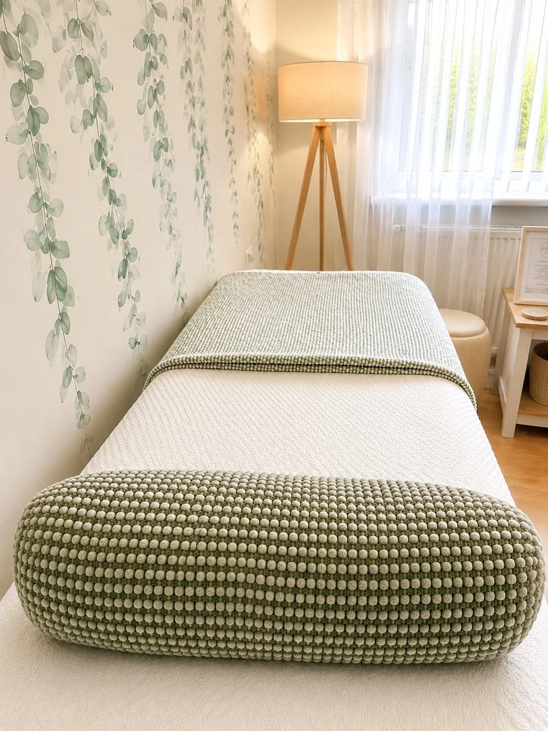 A massage table in a room with light green and white leaf-patterned wallpaper, a wooden floor, a tall floor lamp, a window with sheer curtains, and a small side table with framed certificate and decorations.