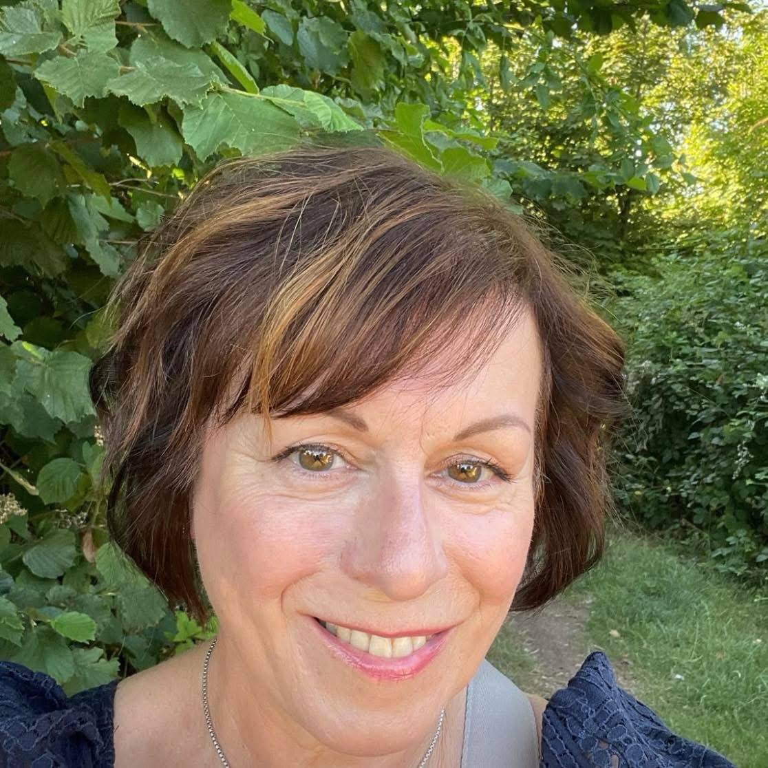 A woman with short brown hair and hazel eyes smiling outdoors with green foliage in the background.
