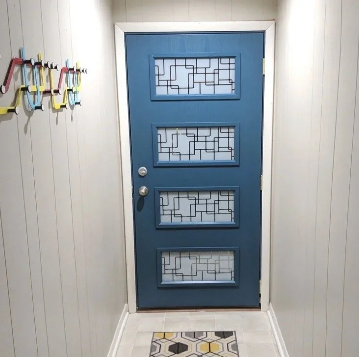 Blue front door with four glass panels featuring geometric patterns, located in a narrow hallway with white paneled walls and a geometric-patterned rug on the floor.