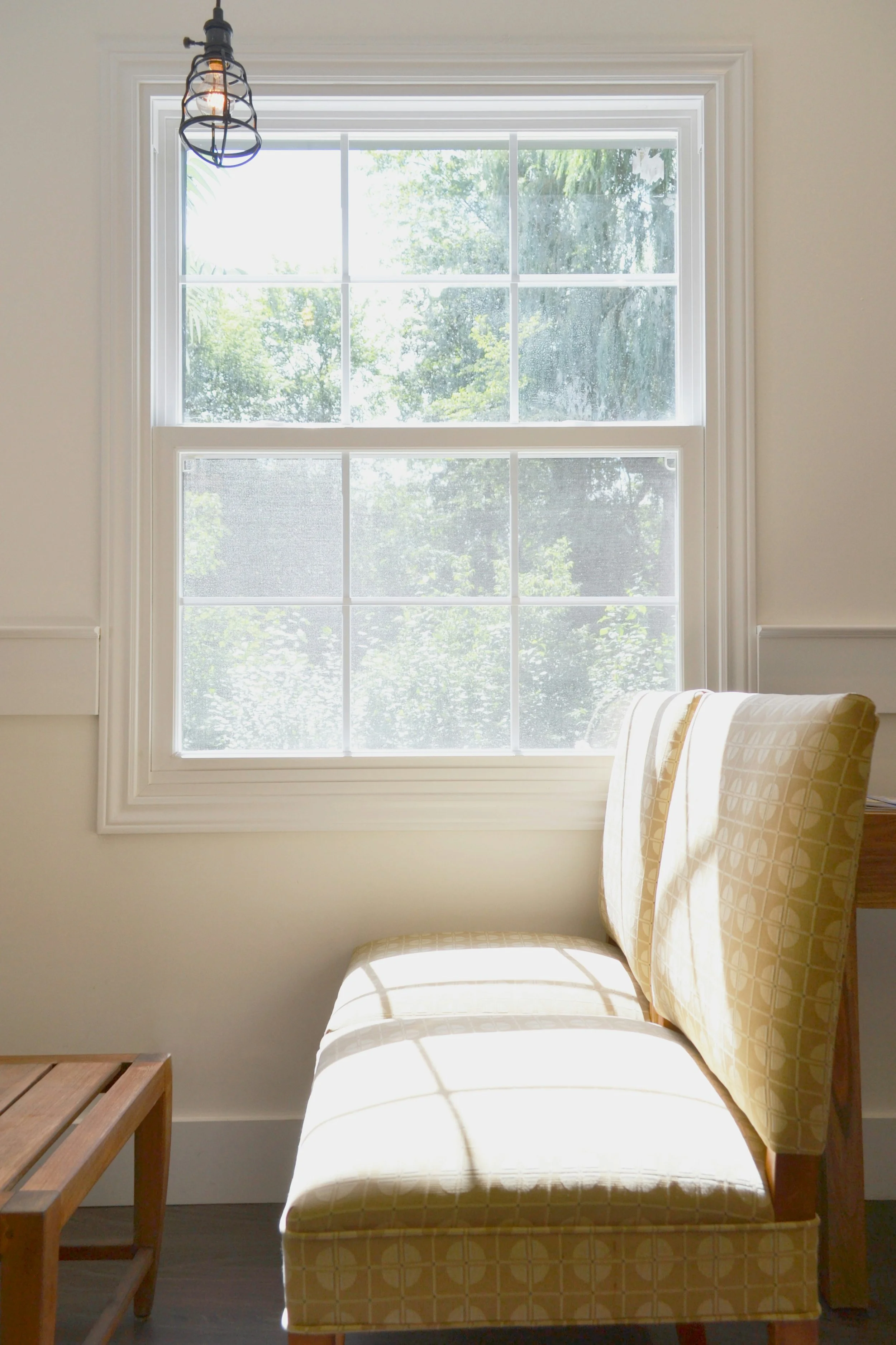 Sunlit room with a large double-pane window showing green trees outside, light-colored walls, a yellow patterned armchair, and a small wooden side table.