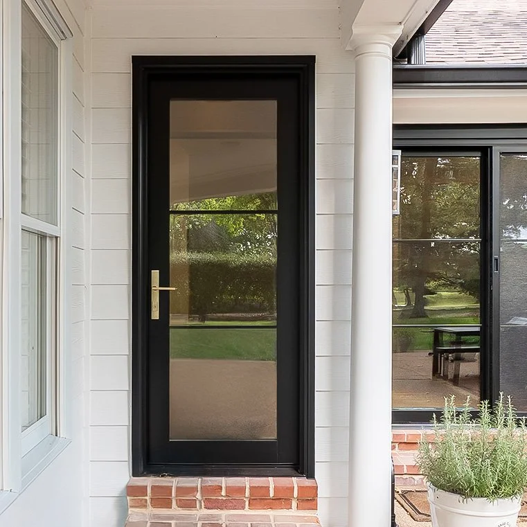 Black-framed door with a glass panel, part of a white house exterior with brick step, white siding, and a large window.