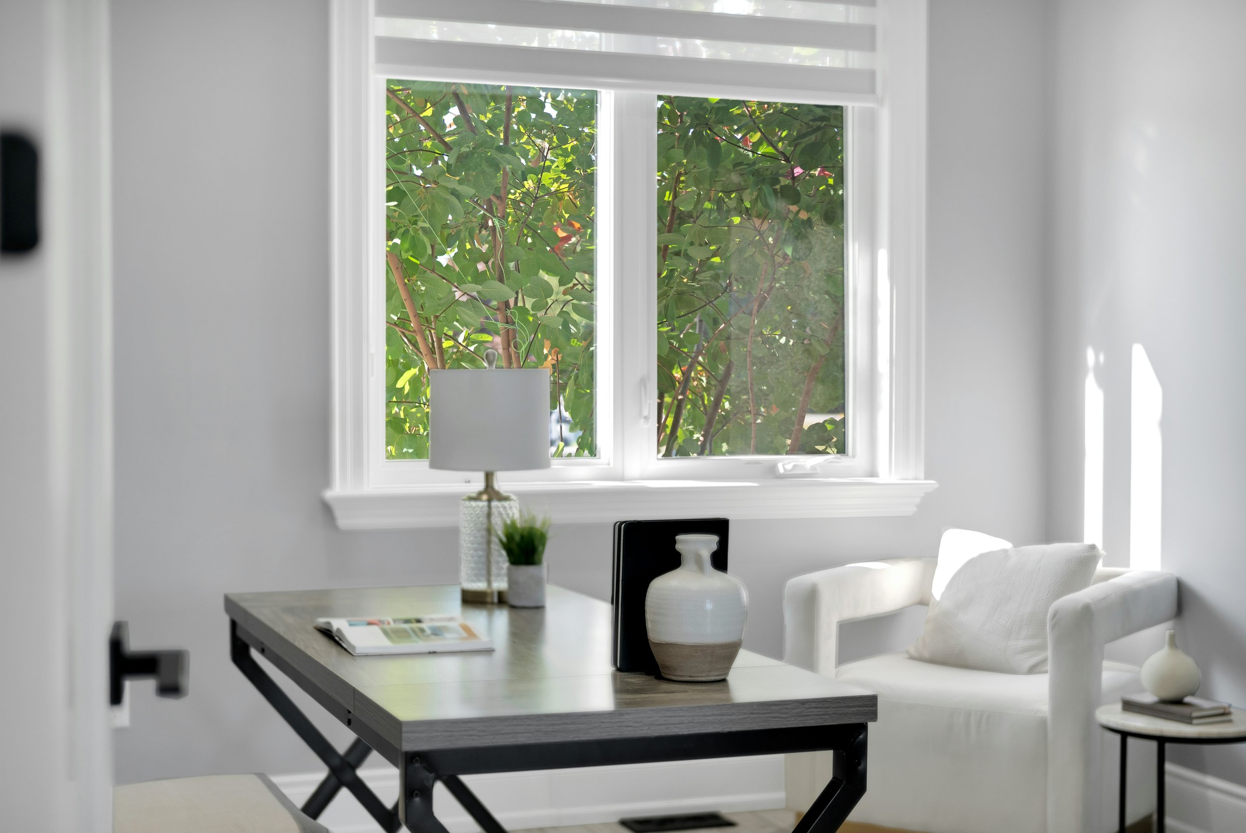 A bright room with a large window showing green leafy trees outside. Inside, there is a white table with a magazine and decorative vases, a table lamp, and a white chair with cushions.