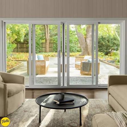 Living room with beige sofas, a round black coffee table with books, and sliding glass doors leading to a backyard patio with outdoor furniture and trees.