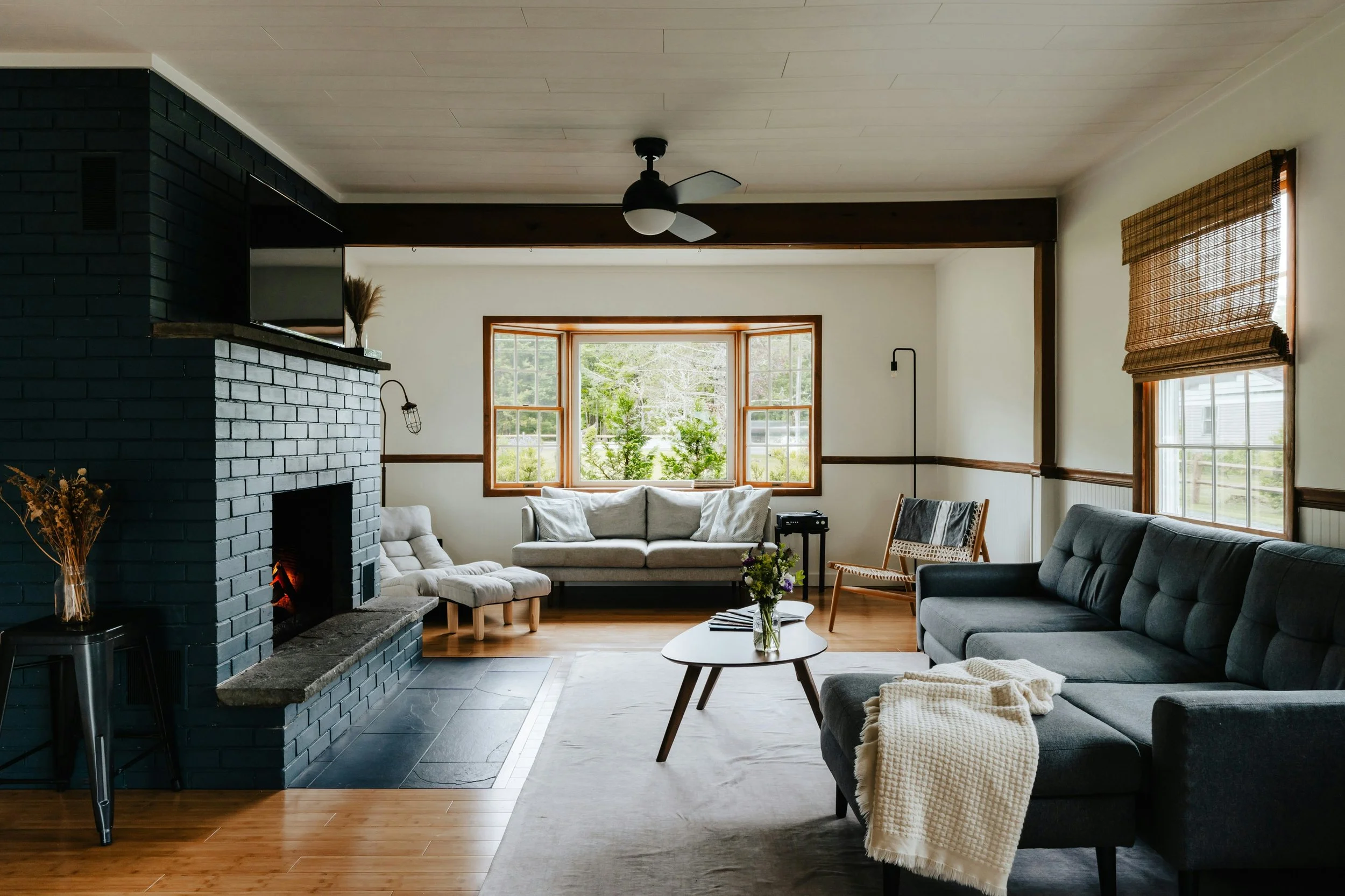 Living room with gray sectional sofa, white armchair, wooden floor, large window with view of greenery, black ceiling fan, and black brick fireplace.