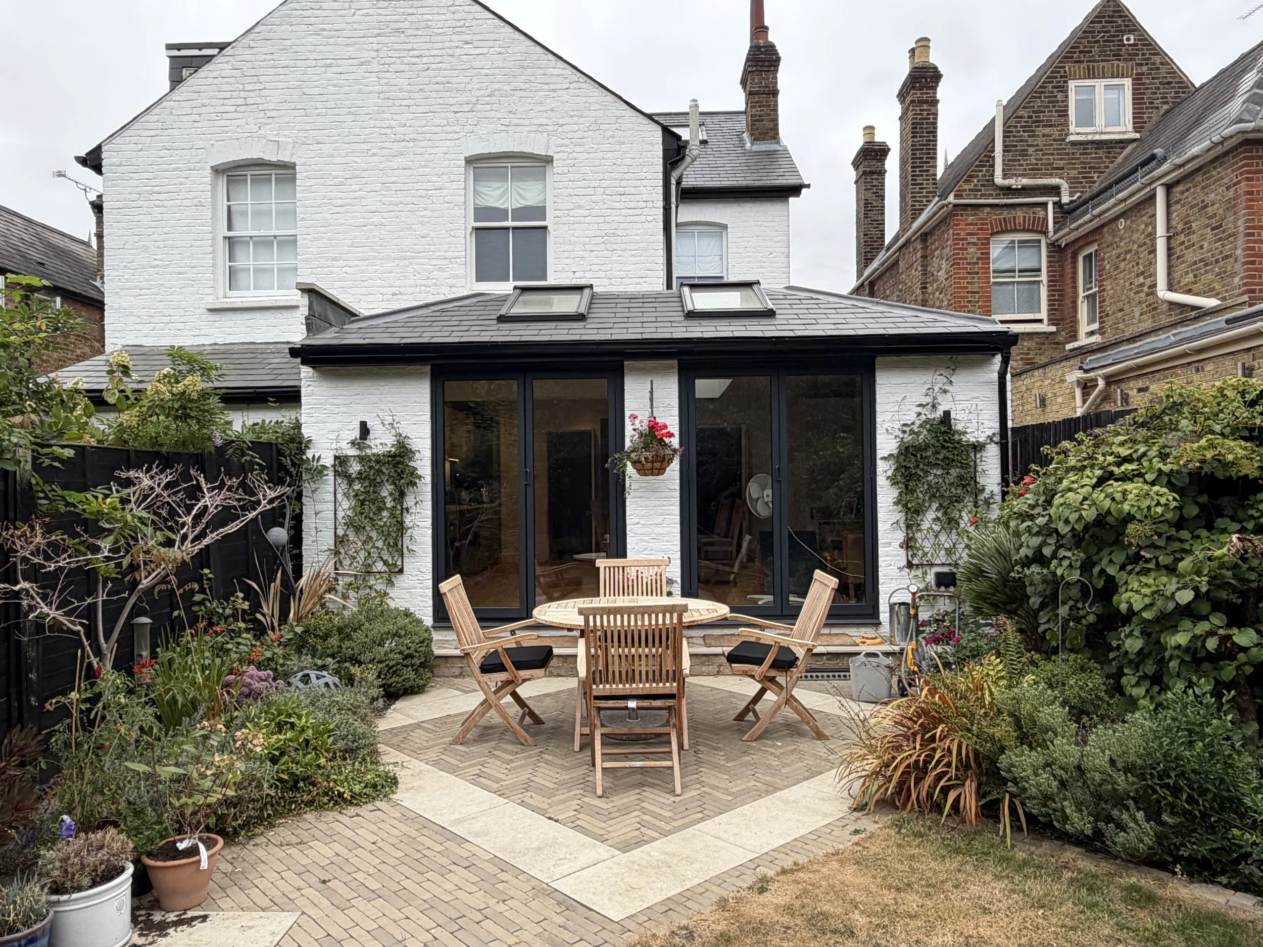 A backyard garden with a round wooden table and four chairs, surrounded by various plants and flowers, with a white brick house in the background featuring large glass doors and windows.