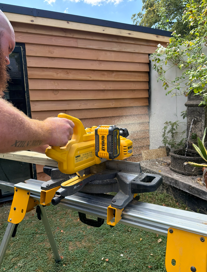 Person using a yellow cordless power saw on a worktable outdoors with a wooden shed and plants in the background.