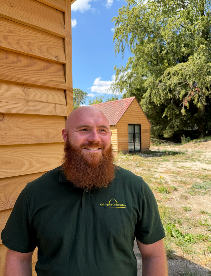 A smiling man with a red beard standing outside near a wooden building, with another similar small wooden building and green trees in the background on a sunny day.