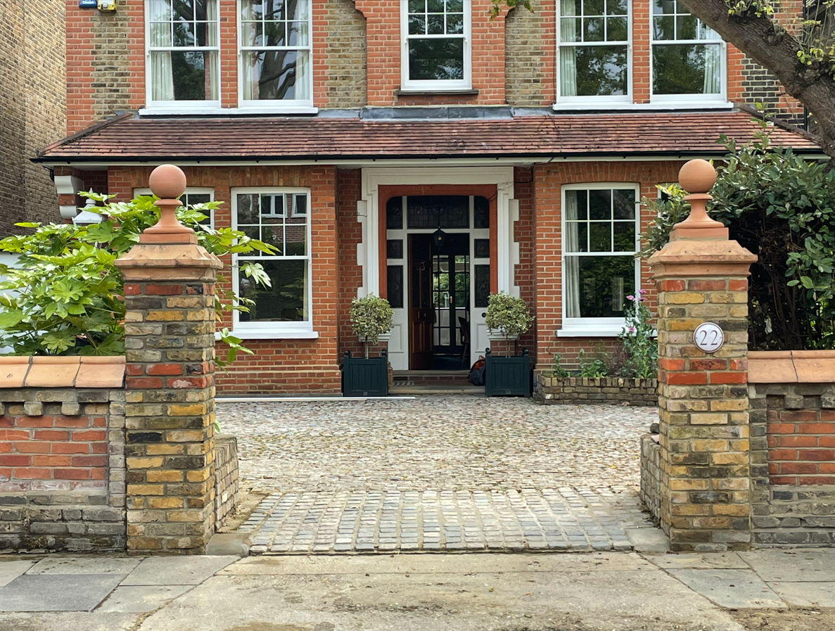 A brick house with white-framed windows and a front door. Two potted plants sit on either side of the door. A brick and stone front wall with pillars and a driveway with cobblestones lead to the entrance. The house number '22' is displayed on a rounded plaque on the right pillar.
