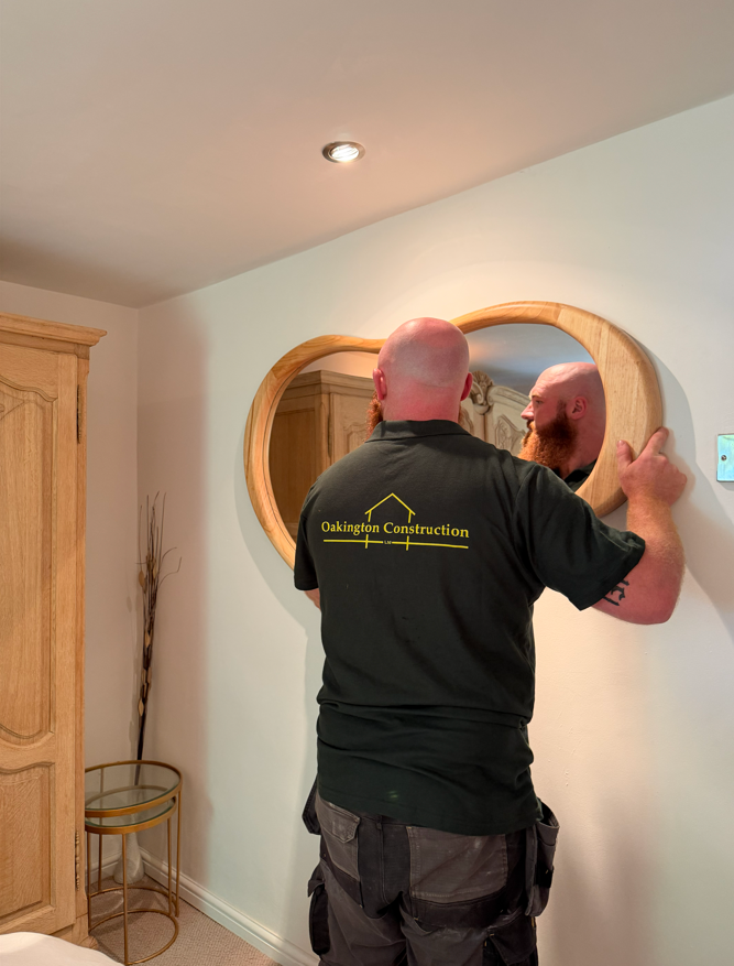 A man with a bald head and red beard looking at himself in a round wooden mirror mounted on the wall. He is wearing a black shirt with the logo 'Oakington Construction' on the back, in a room with light-colored walls and wooden furniture.