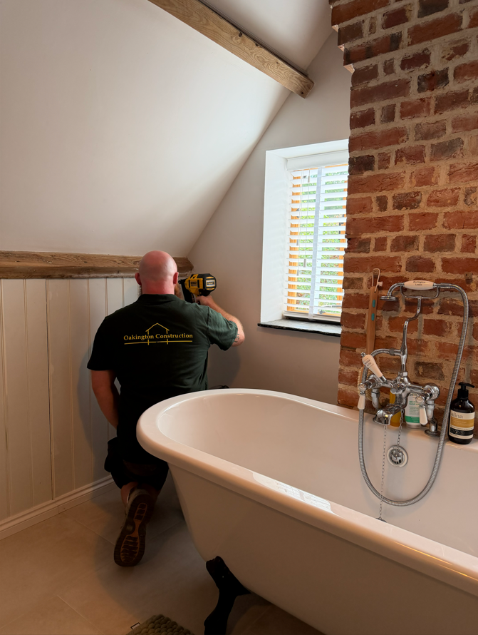 A worker kneeling near a clawfoot bathtub, installing or repairing something using a cordless drill. The worker is wearing a black shirt with the logo 'Oakington Construction'. The room has a slanted ceiling with exposed wooden beam, a brick wall, a window with shutters, and bathroom essentials on a faucet.