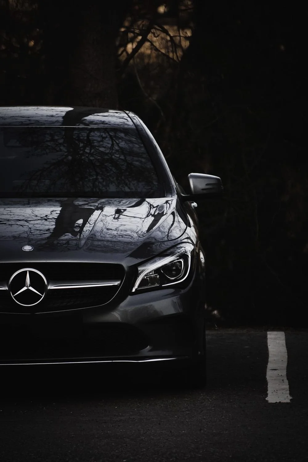 Close-up of the front of a black Mercedes-Benz car, parked on a dark road with a white parking line, reflections of trees visible on the car's hood and windshield, dark background with trees.