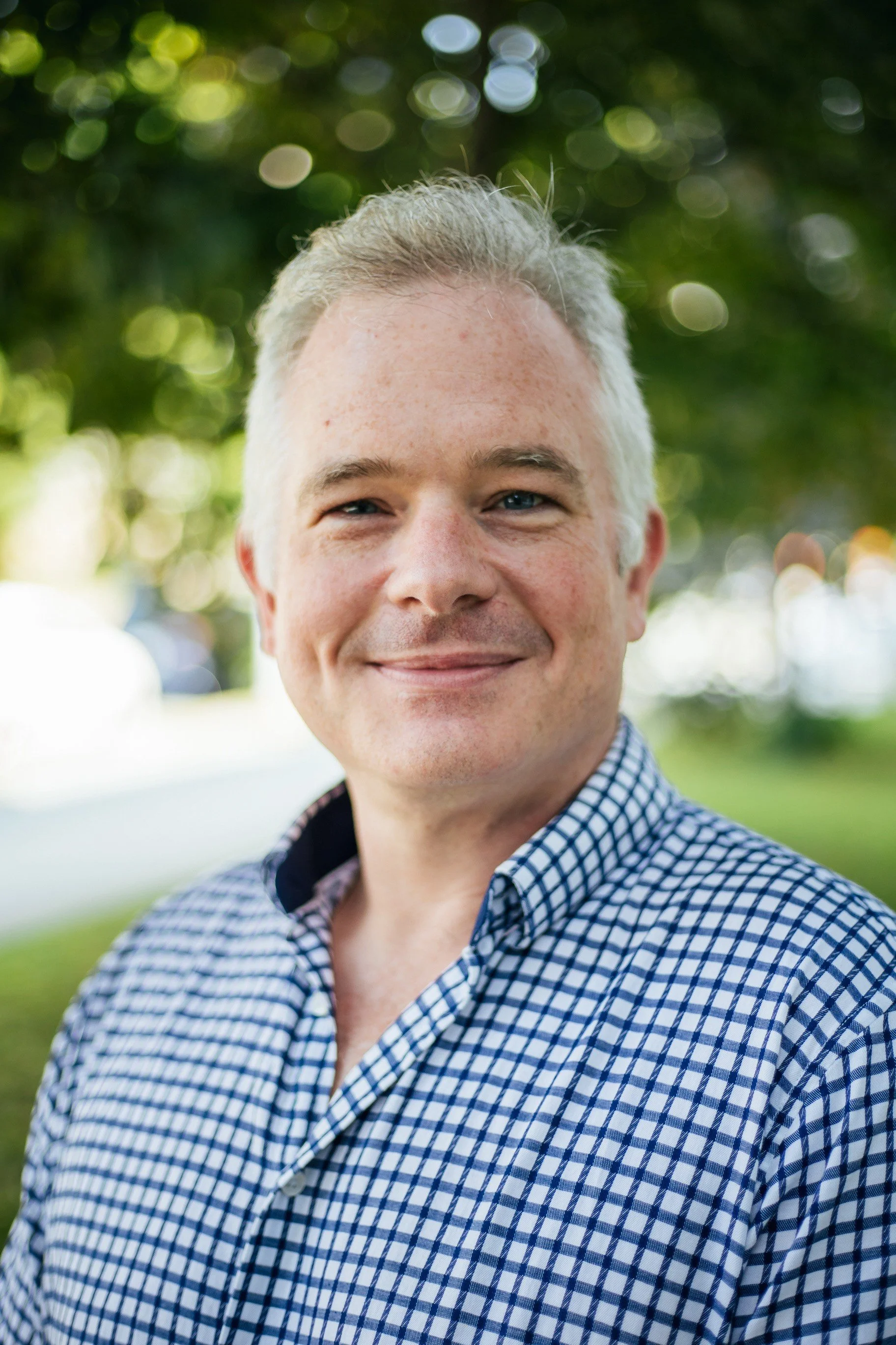 Close-up of a smiling middle-aged man with gray hair, blue eyes, wearing a checkered button-up shirt, outdoors with green trees and bokeh sunlight in the background.