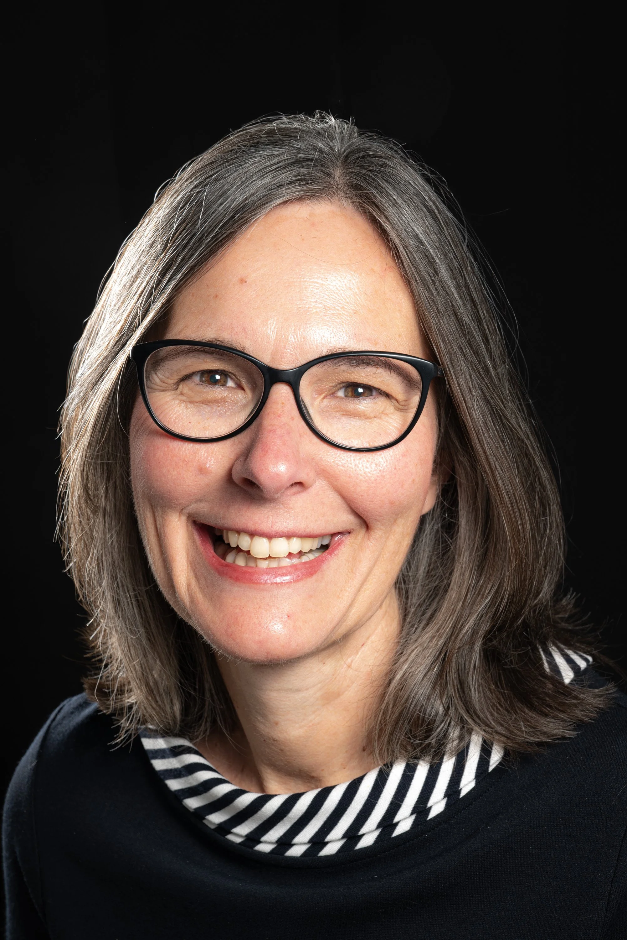Close-up of a smiling woman with gray hair, wearing glasses, a black and white striped shirt, and a black sweater, against a black background.