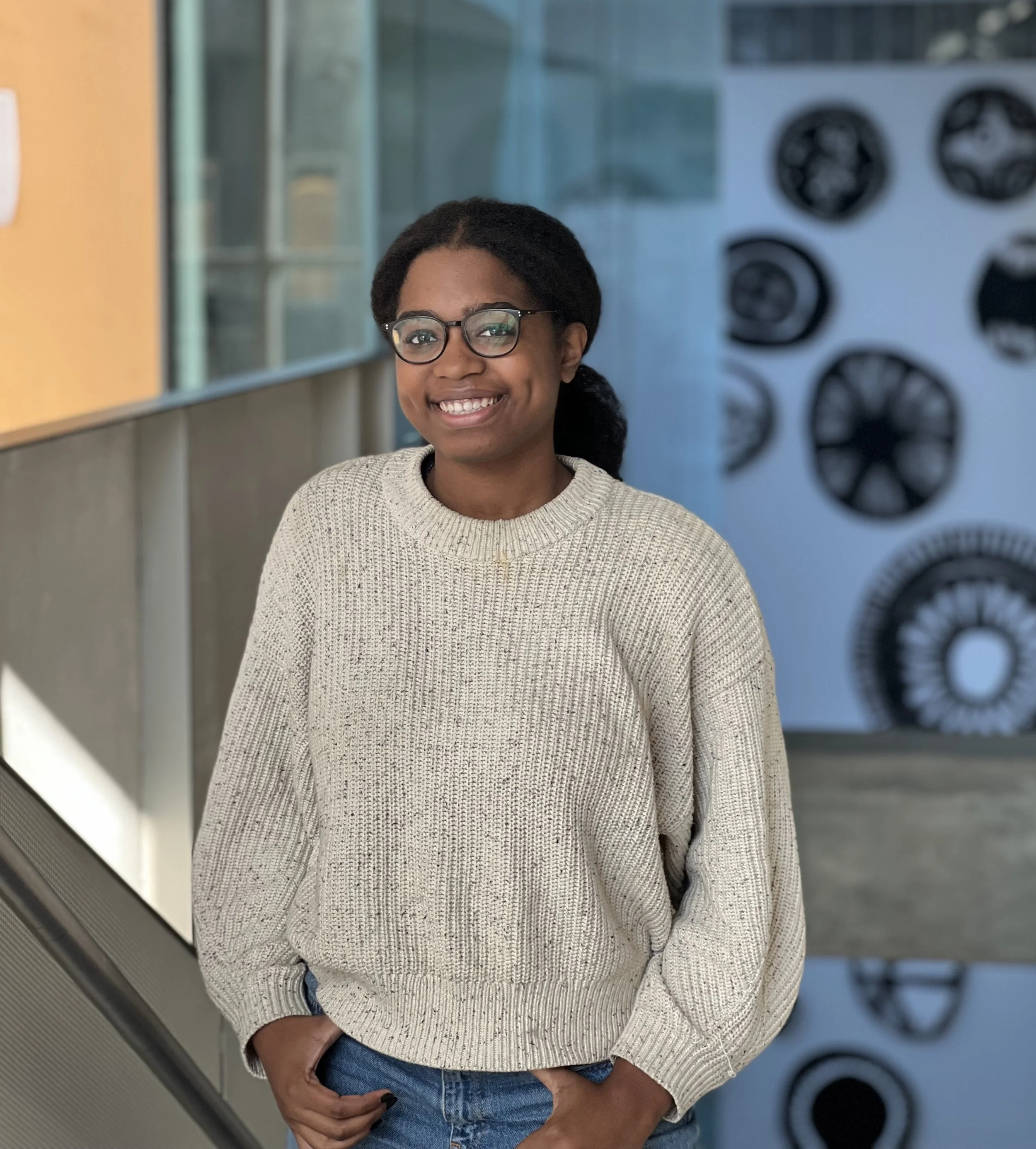 A woman with dark curly hair, glasses, and a big smile standing indoors in front of a wall decorated with circular objects, wearing a beige chunky knit sweater and blue jeans.