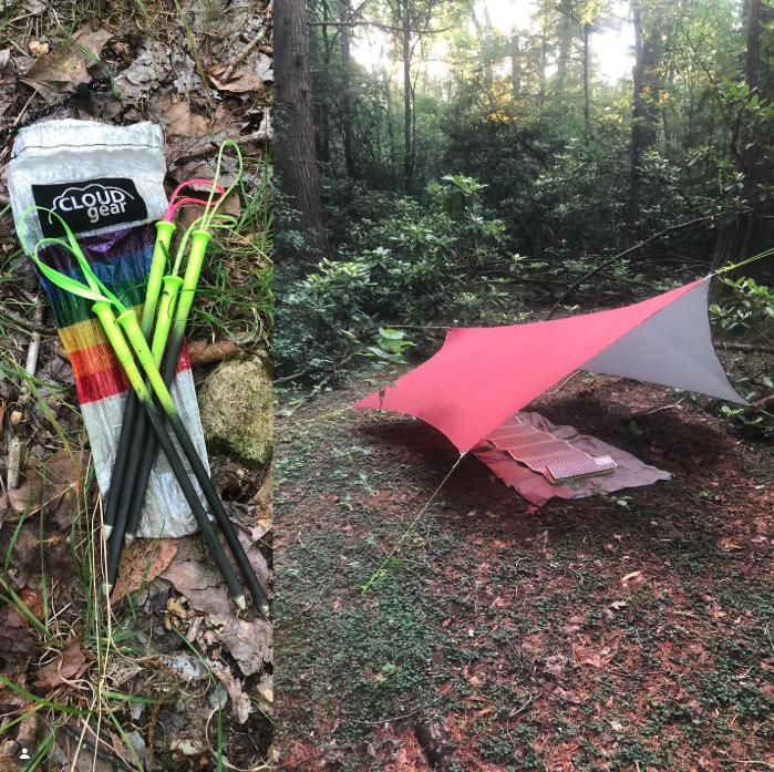 Camping setup in a forest with a pink and gray tarp shelter and a pack of trekking poles laid on the ground surrounded by leaves and grass.