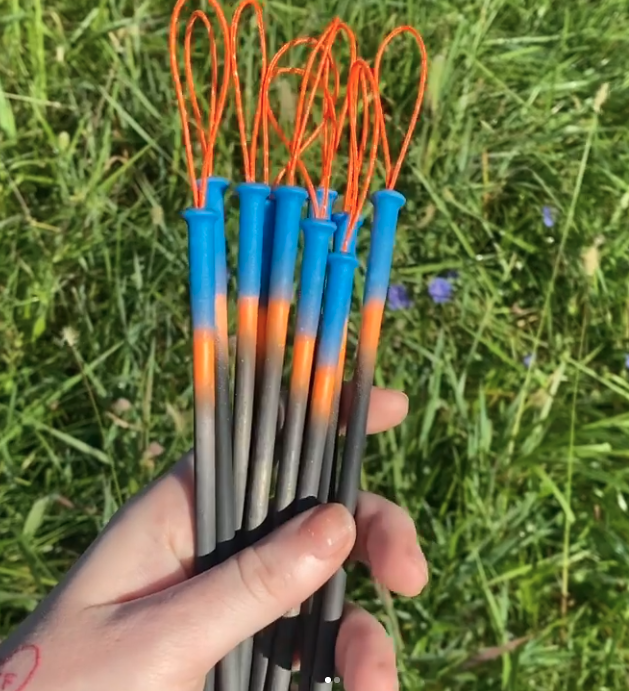 A hand holds a bunch of colorful fireworks with blue, orange, gray, and black handles and orange loops at the tips, against a background of green grass and small purple flowers.
