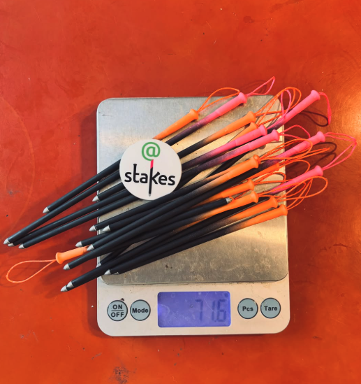 A digital kitchen scale weighs ten black golf tees with orange and pink tops. A sticker with the '@' symbol and the word 'stikes' is placed on the tees.