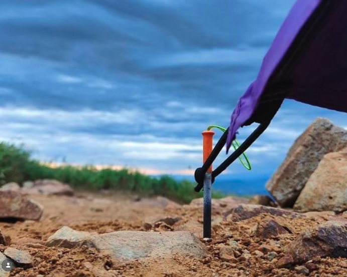 Close-up of a purple backpack with a hydration tube on rocky ground outdoors, with a cloudy sky and distant greenery in the background.