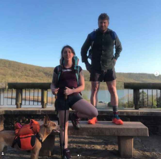Two hikers, one woman and one man, and a dog on a trail overlooking a river and hills, during daytime with clear skies.