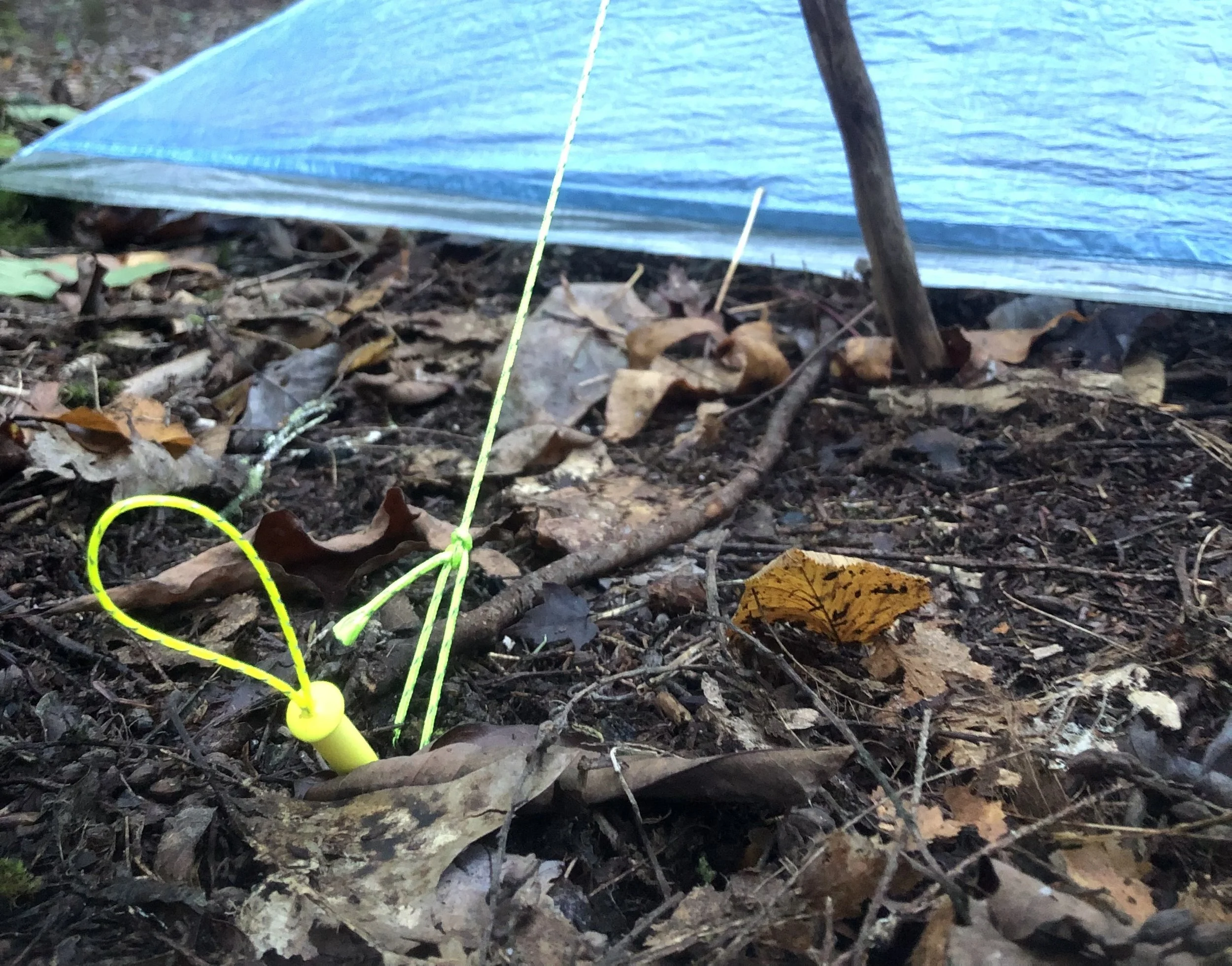 Close-up of a small yellow fishing bobber with a bright yellow string, lying on a bed of dry leaves and twigs on the forest floor, with a blue tarp and tree trunk in the background.