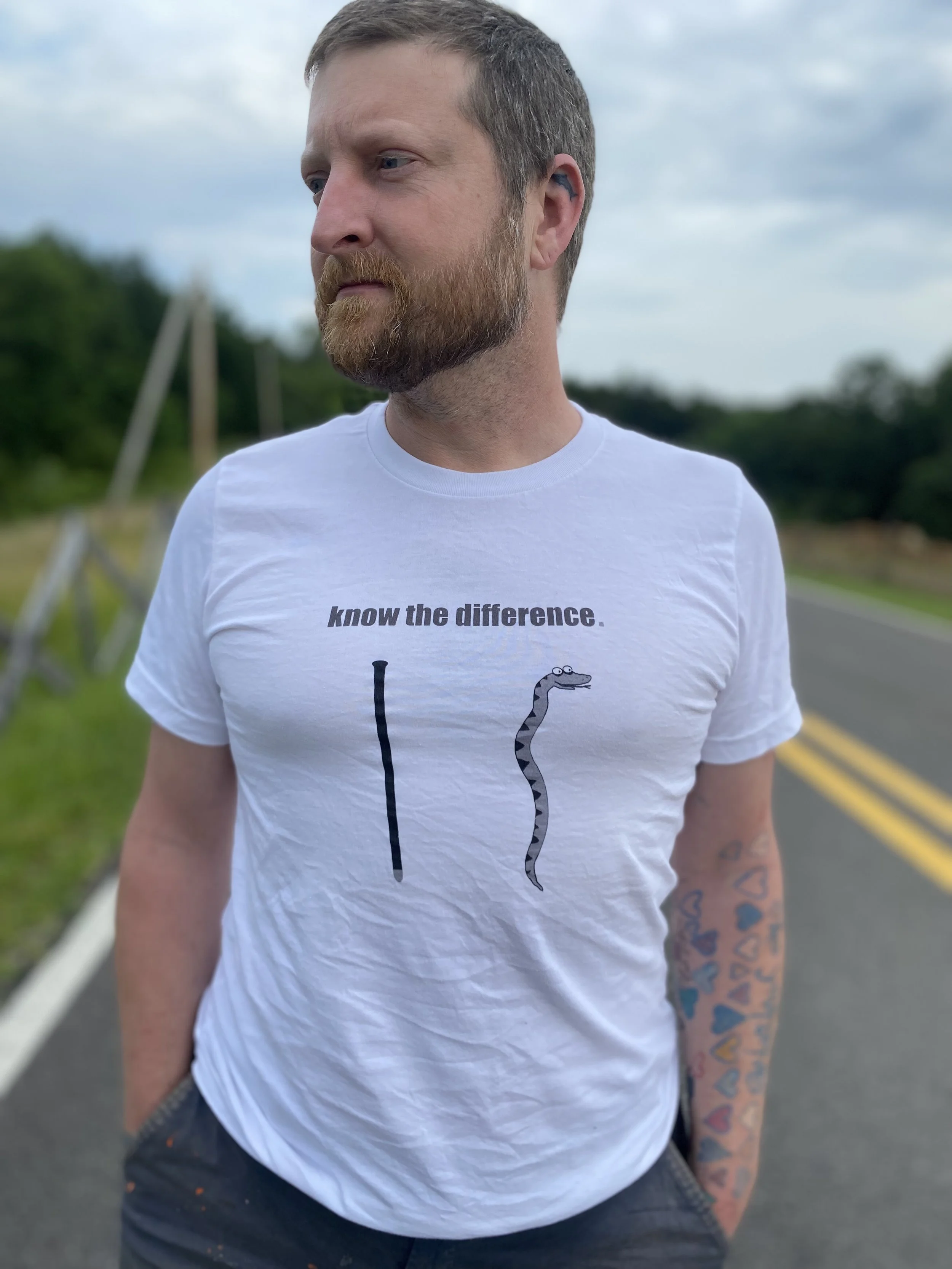 A man with a beard wearing a white T-shirt with a snake illustration and the text "know the difference." standing on a rural road with trees and utility poles in the background.