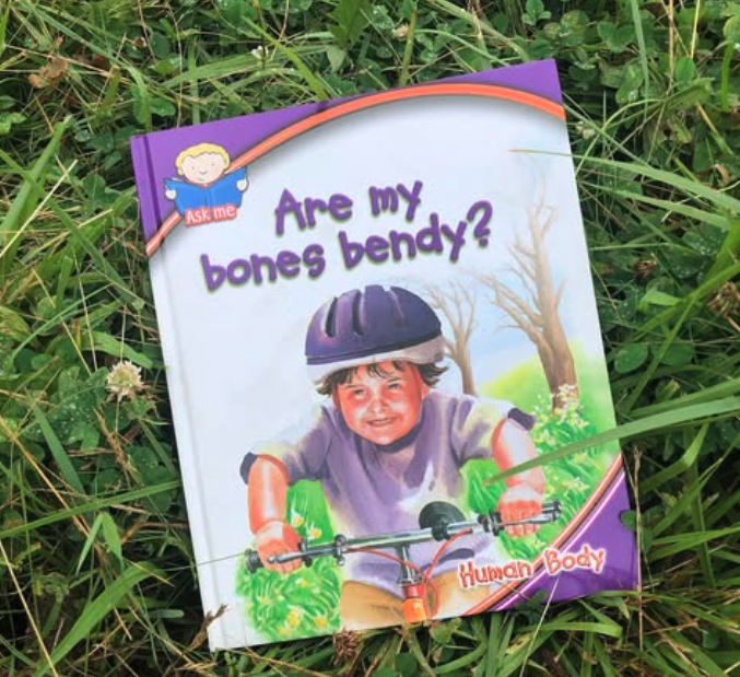 A children's book titled "Are my bones bendy?" lying in grass. The cover features a child wearing a bicycle helmet, smiling while riding a bike, with trees in the background.
