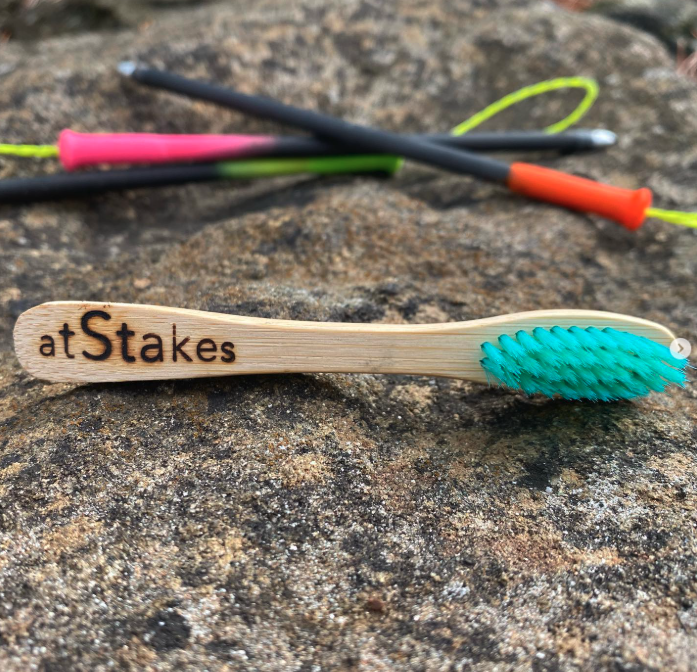 A wooden toothbrush with the words "at Stakes" engraved on the handle, and a green and blue toothbrush head, resting on a rough, textured surface with three colorful pens or markers in the background.