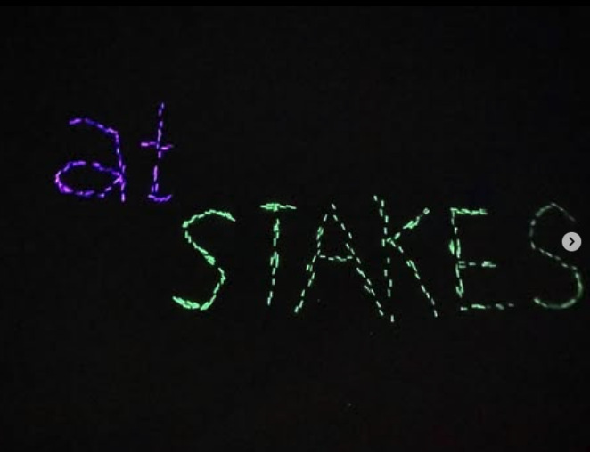 String of lights in the dark spelling 'at STAKES' with 'at' in purple and 'STAKES' in green.
