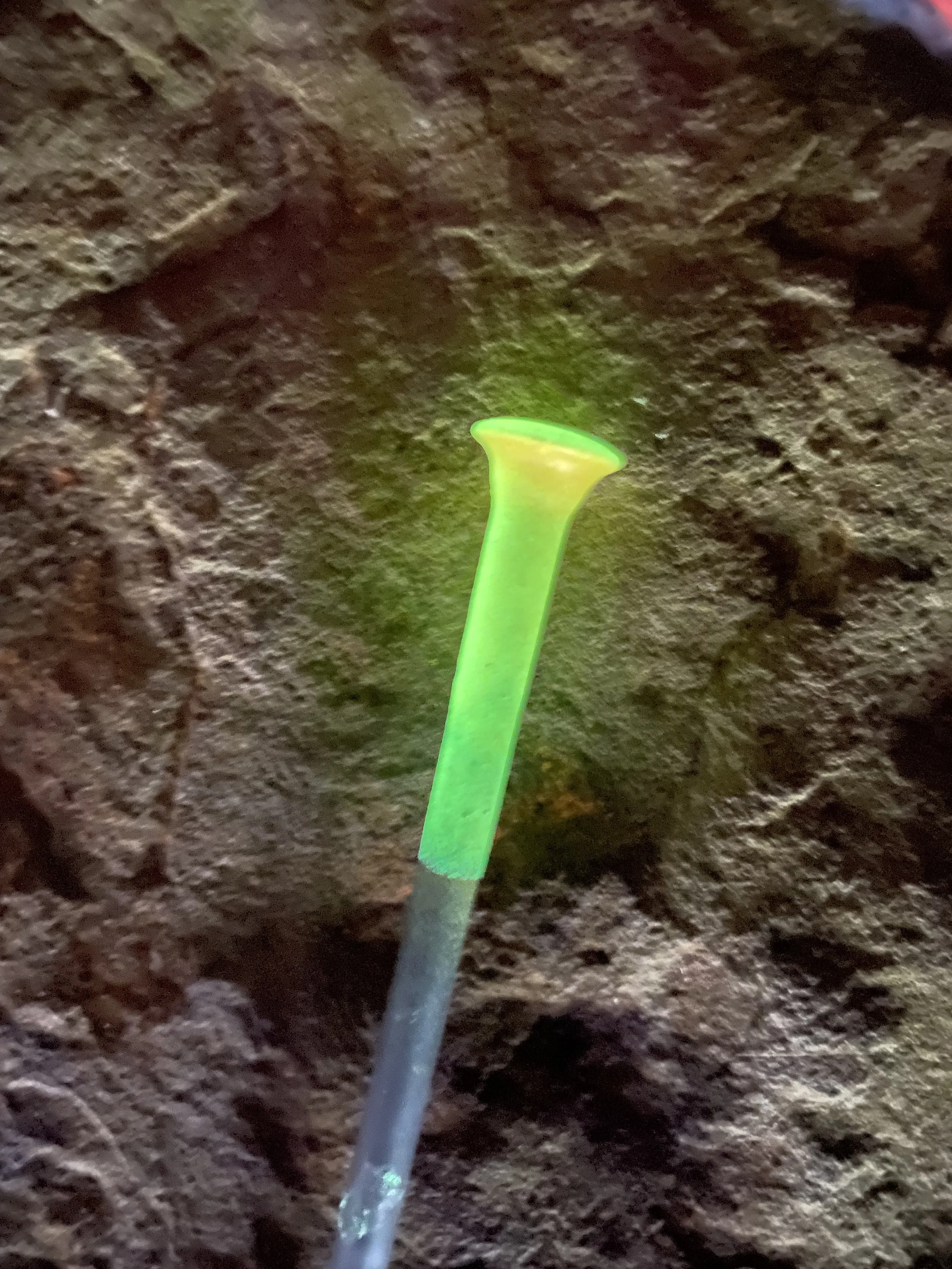 A small green and yellow plastic shovel sticking into a dirt surface, with a rough, uneven, brown texture.