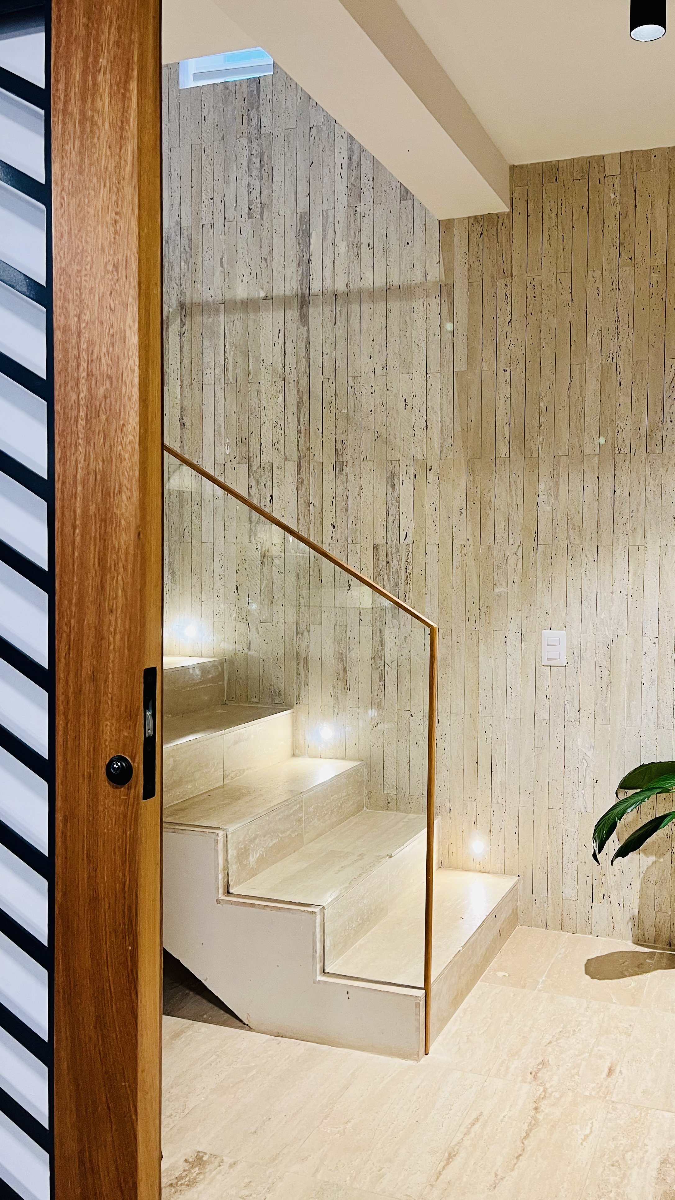 Completed interior staircase designed by Yamilis de la Rosa and built by Constructora Caña Roja in Casa de Campo, Dominican Republic, featuring natural stone, integrated step lighting, glass railing, and wood detailing.