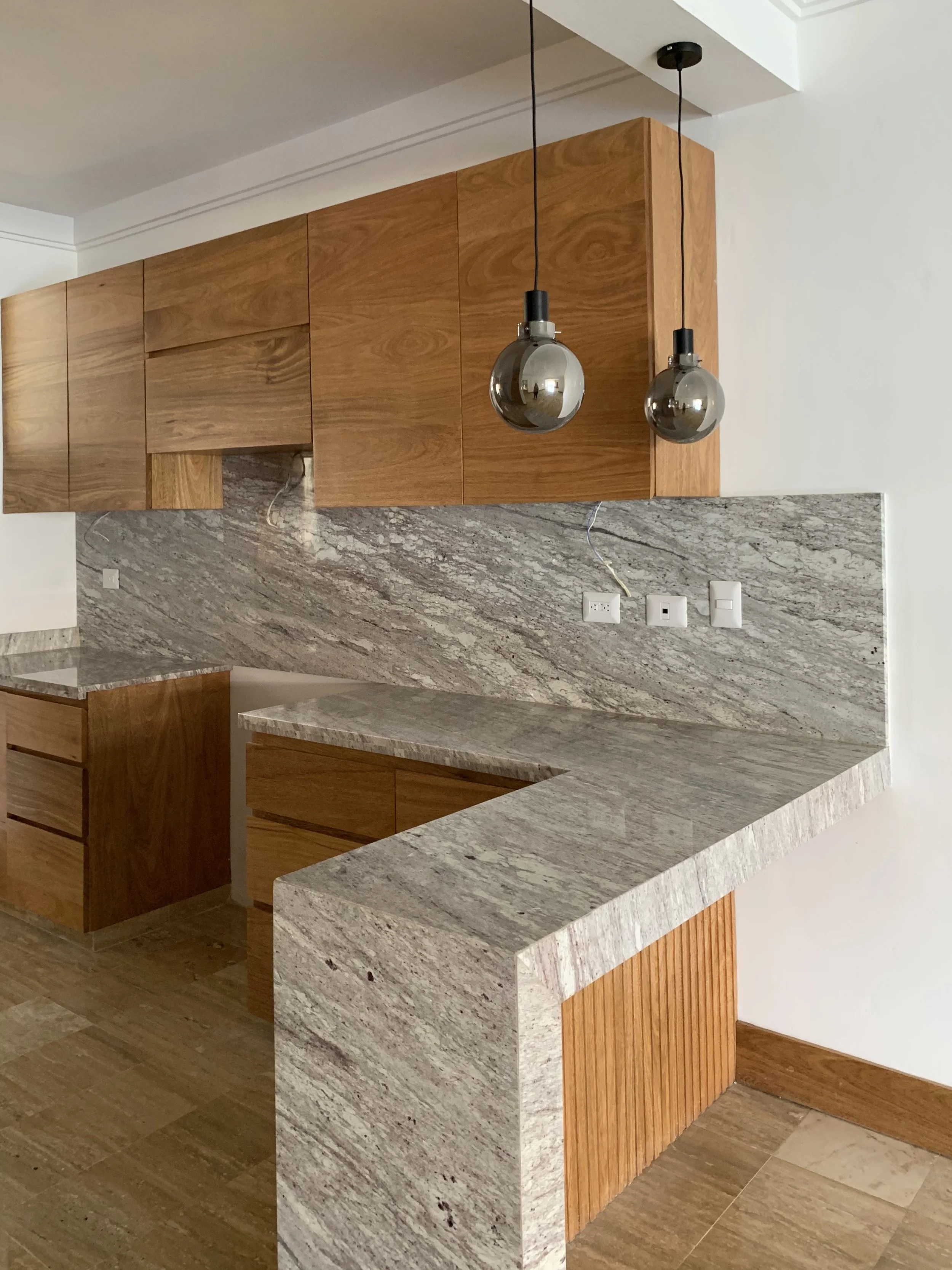 Completed residential kitchen designed by Yamilis de la Rosa and built by Constructora Caña Roja in Casa de Campo, Dominican Republic, featuring custom wood cabinetry, natural stone surfaces, and pendant lighting.