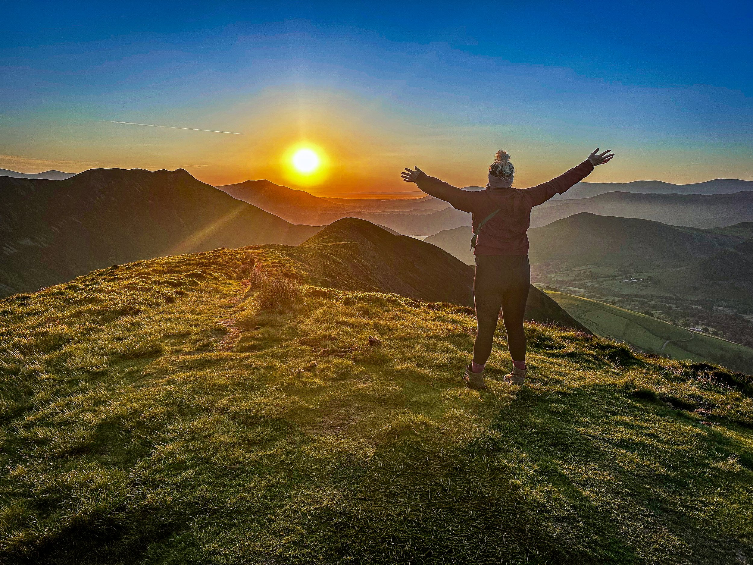 A person standing on a grassy mountain ridge with arms outstretched, facing a sunrise over distant mountain ranges on a clear day.