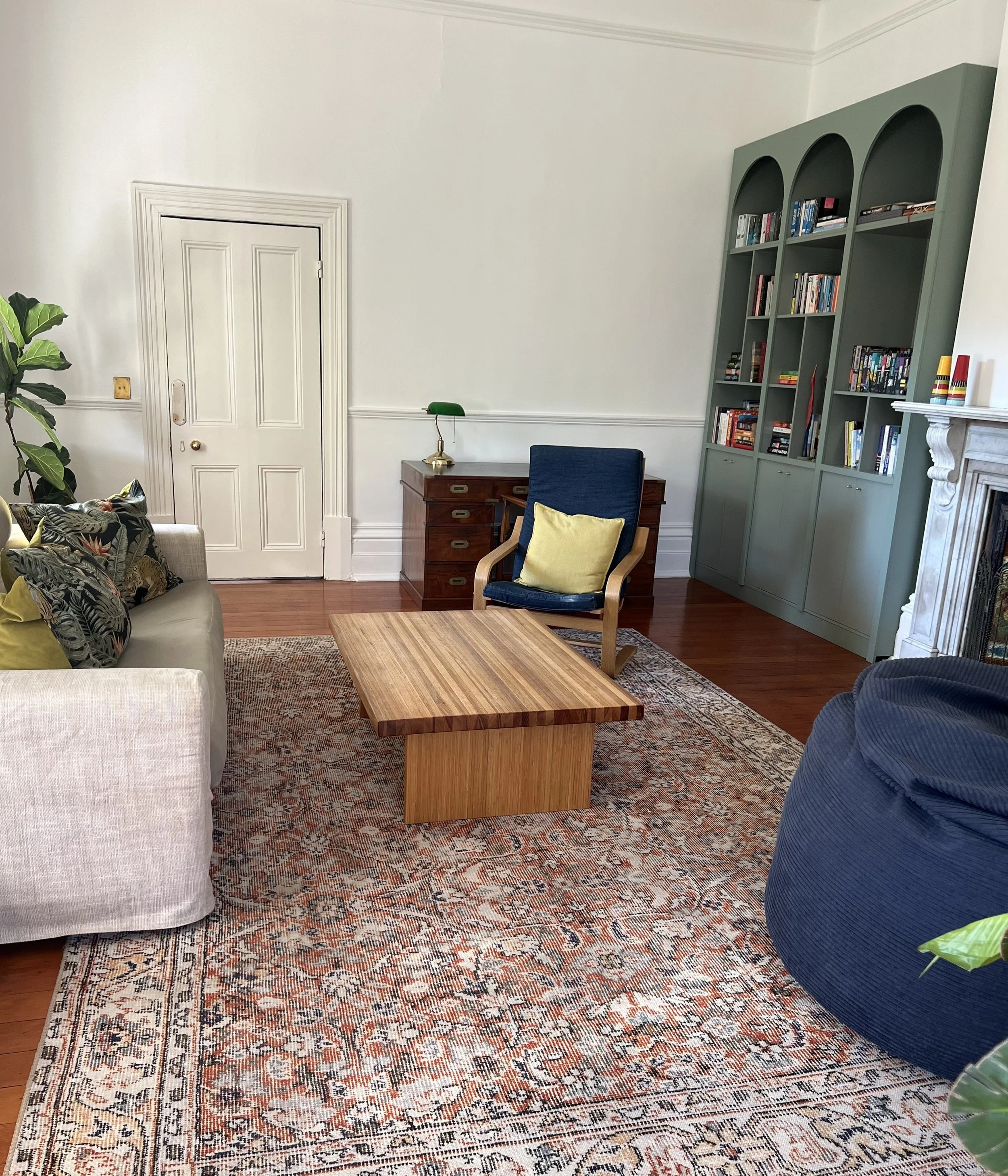 Interior room with white walls, a patterned rug, a white sofa with patterned pillows, a wooden coffee table, a white fireplace, and a tall green bookcase filled with books. There is a wooden desk with a green banker lamp and a blue chair with a yellow pillow.